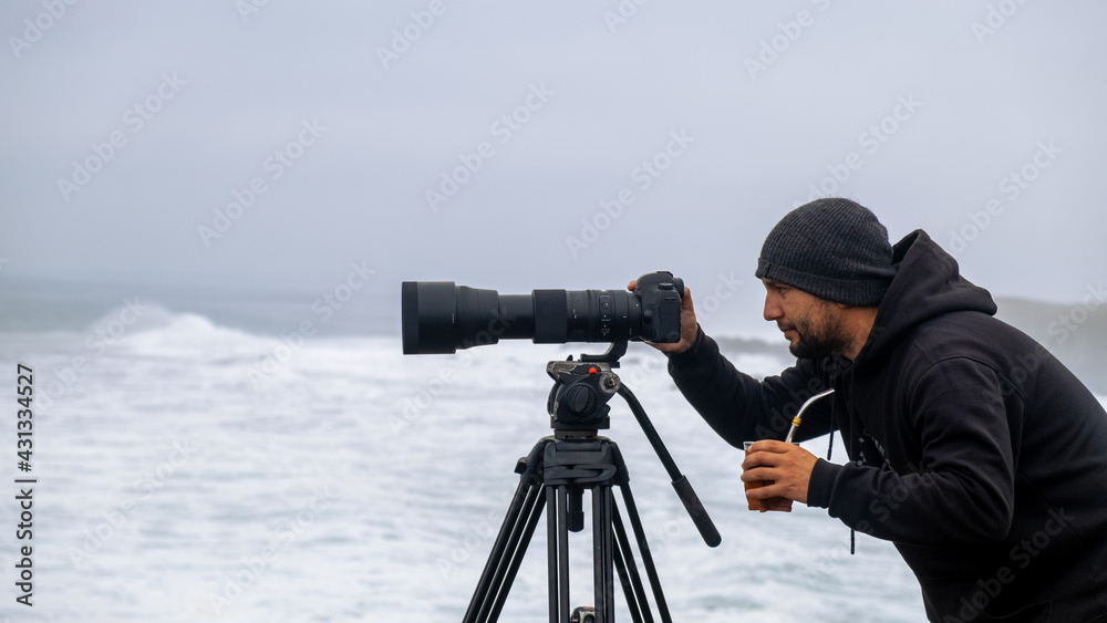 Surf photographer taking photos of the sea and surfers on a cloudy day with his camera and telephoto
and mate argentine drink 