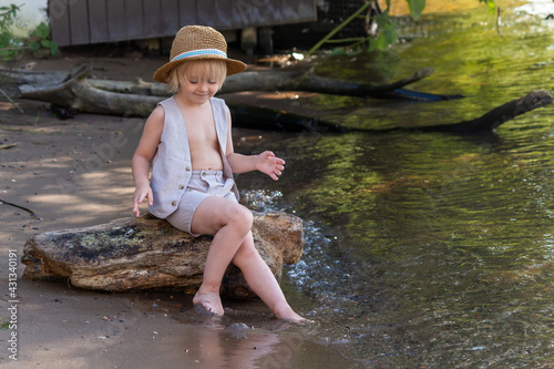 Fototapeta Naklejka Na Ścianę i Meble -  Little cute caucasian blond boy wearing straw hat sits on the tree log by the river. Copy space for your text. Summer kids fun theme.
