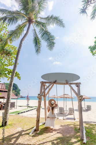 Back View of Travel Woman in White Dress and Straw Hat Sitting on Wooden Swing, Tropical Palms and Sandy Beach with Blue Sea on Background. Female Tourist Leisure in Resort on Phuket Island, Thailand