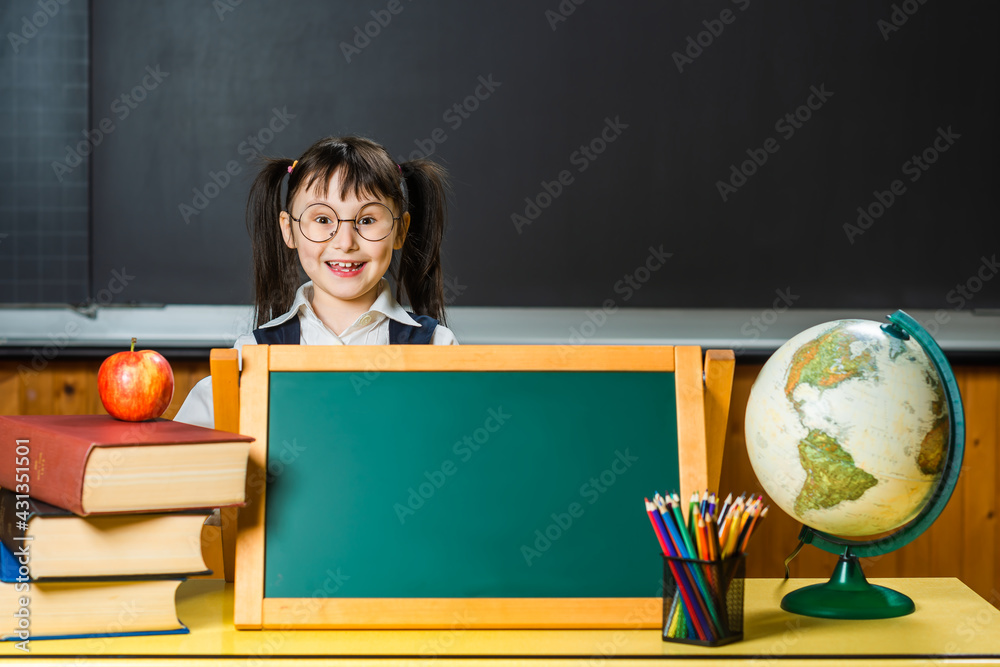 Little elementary school girl in classroom interior. Books, globe, apple, pencils, marker board, chalk board. Lots of space for text, messages, advertising. Little elementary school girl in classroom interior. Books, globe, apple, pencils, marker board, chalk board. Lots of space for text, messages, advertising.