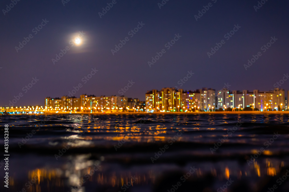 Residential buildings with lanterns stand on the embankment of the river. Full moon in the night sky. Panorama of the night city. Close up view from the shore level.