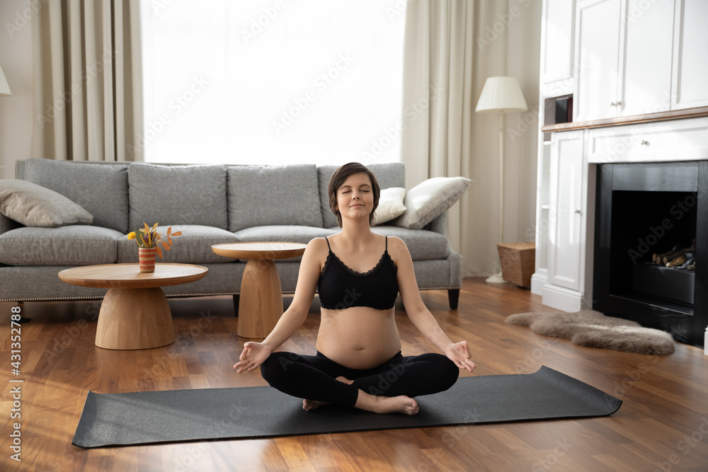 Calm young Caucasian pregnant woman sit on mat in living room at home practice yoga. Happy millennial female follow healthy lifestyle during pregnancy, breathe fresh ventilated air. Sports concept.