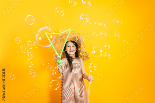 Little happy girl has fun blowing huge soap bubbles. Yellow bright wall background. Positive children's emotions.