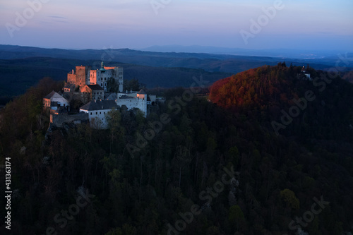 Buchlov Castle. Aerial view on monumental castle in Romanesque Gothic style, standing on a wooded hill against Saint Barbara’s Chapel on the hill in background. Castle in the dark. Czech republic