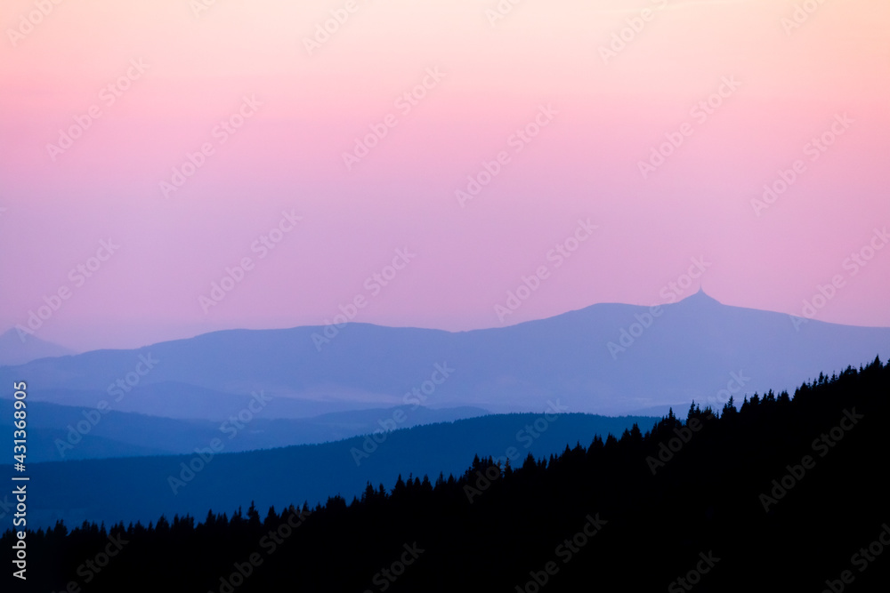 Colorful sunset in Czech mountains. Blue mountain ranges layers against pink to orange evening sky in the Krkonose national park.