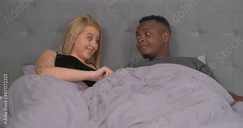 Multiethnic couple lying in bed. Caucasian woman lifting blanket and amazed of male's erection. Sexual health and relationship