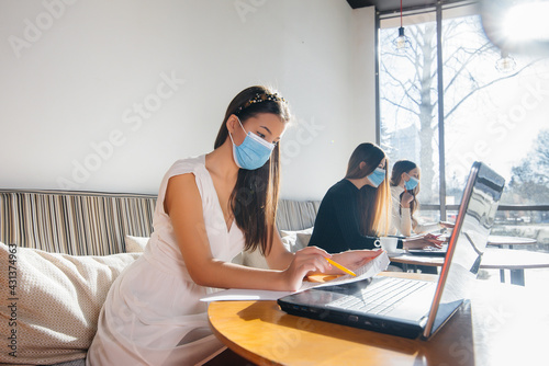 A group of masked girls keep a social distance in a cafe when working on laptops.