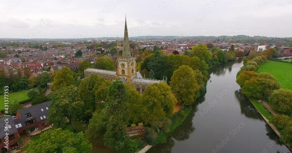 Aerial Approaching The Medieval Gothic Holy Trinity Church On The River Avon, Surrounded By Lush Green Trees And The Village Homes In The Background - Stratford-upon-Avon, United Kingdom Aerial Approaching The Medieval Gothic Holy Trinity Church On The River Avon, Surrounded By Lush Green Trees And The Village Homes In The Background - Stratford-upon-Avon, United Kingdom