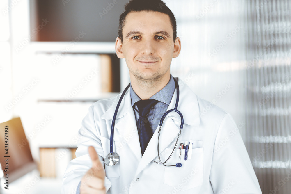 Male doctor standing with thumbs up sign in clinic near his working place. Perfect medical service in hospital. Medicine and healthcare concept Male doctor standing with thumbs up sign in clinic near his working place. Perfect medical service in hospital. Medicine and healthcare concept