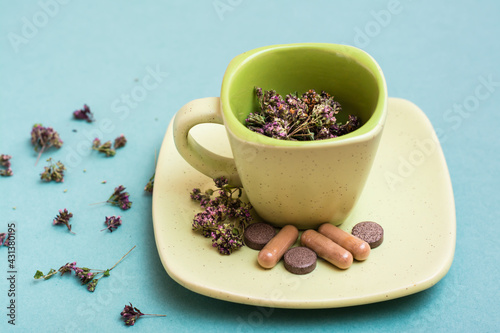A cup with dried oregano and herbal medicinal capsules and pills on a saucer on a green background. Alternative medicine