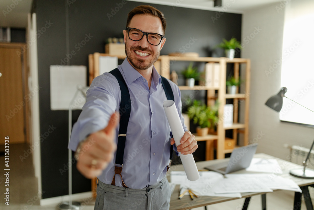 Smiling young architect posing in his office, holding blueprint and showing thumb up. Smiling young architect posing in his office, holding blueprint and showing thumb up.