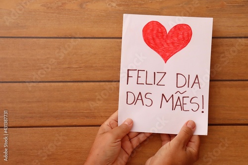 Mom holding Feliz Dia das Mães (Happy Mother's Day, in portuguese) gift card on wooden background, with copy space. 
