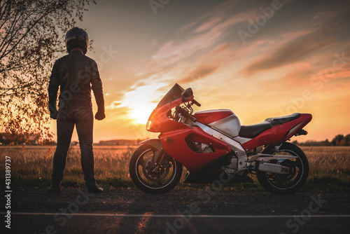 A motorbiker is standing on the road in the sunset rays and looking far.