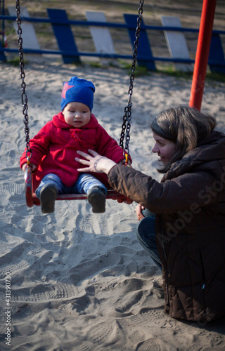 little girl in a red coat and blue hat on a swing with mom in the park