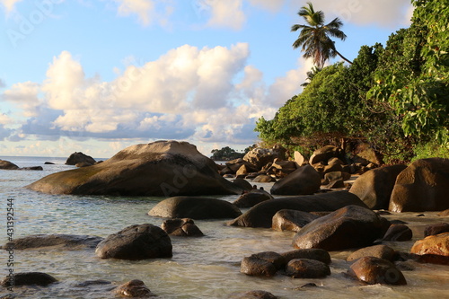 Image of a picturesque sea shore illuminated by sunlight with large smooth stones in the water with trees on the steep slope of the coastline with a blue sky and clouds on the horizon.Tropical Island