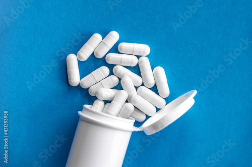 White cylindrical capsules on a matte blue background fall out of white tube. View from above. Medical concept of treatment, prevention and side effects.
