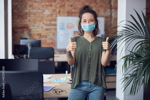 Cheerful creative woman wearing protective mask smiling at camera, showing thums up while standing in modern green office on a daytime photo