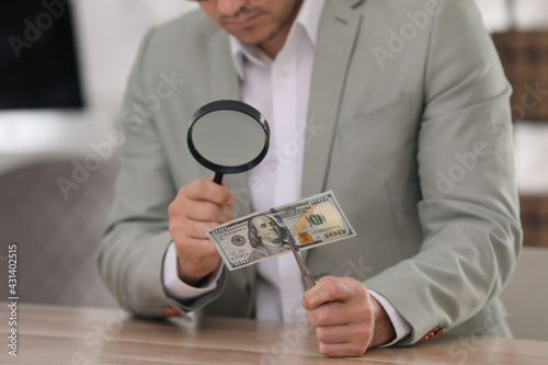 Expert authenticating 100 dollar banknote with magnifying glass at table in office, closeup. Fake money concept
