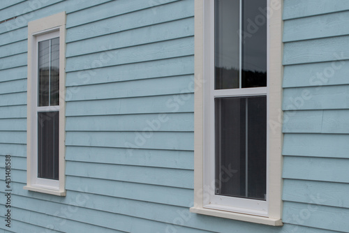 The exterior wall of a baby blue country style house with narrow wood cape cod siding. There are two small double hung windows with white trim. Lace curtains are hanging in the closed glass windows.