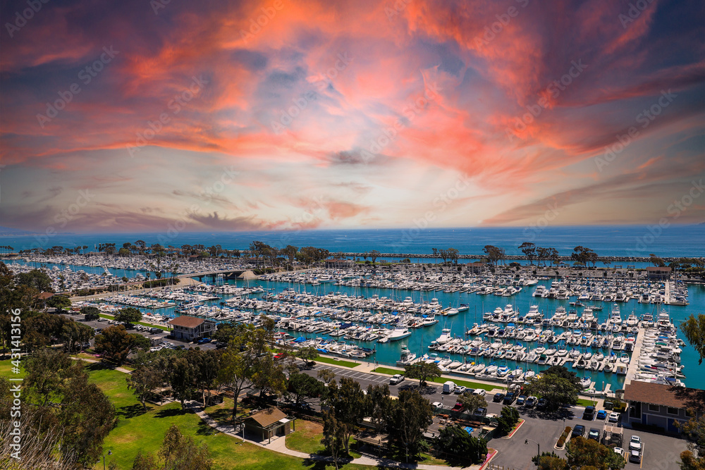 a stunning shot of the boats and yacht docked and sailing Dana Point Harbor with blue ocean water, lush green plants and trees on the hillside and powerful clouds at sunset in Dana Point California - obrazy, fototapety, plakaty 