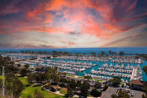 a stunning shot of the boats and yacht docked and sailing Dana Point Harbor with blue ocean water, lush green plants and trees on the hillside and powerful clouds at sunset in Dana Point California #431413156