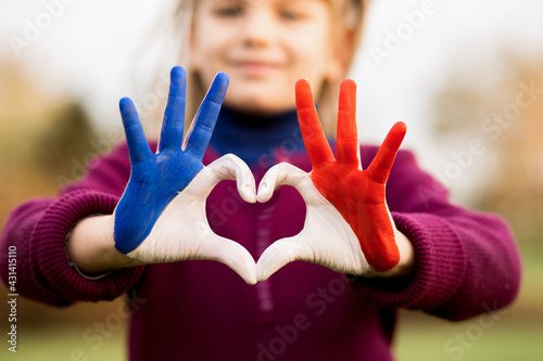 Love and happiness concept. Cute child forming heart gesture with hands outdoors on nature sunset bokeh background. Heart shape of kids hand painted in france flag colors, kids body language