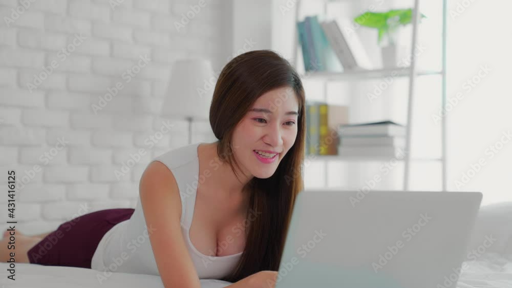 Smiling Asian woman resting in bed. Girl look at laptop screen enjoy pleasant personal conversation using video conference application.Technology help life easy remote communication from home concept Smiling Asian woman resting in bed. Girl look at laptop screen enjoy pleasant personal conversation using video conference application.Technology help life easy remote communication from home concept