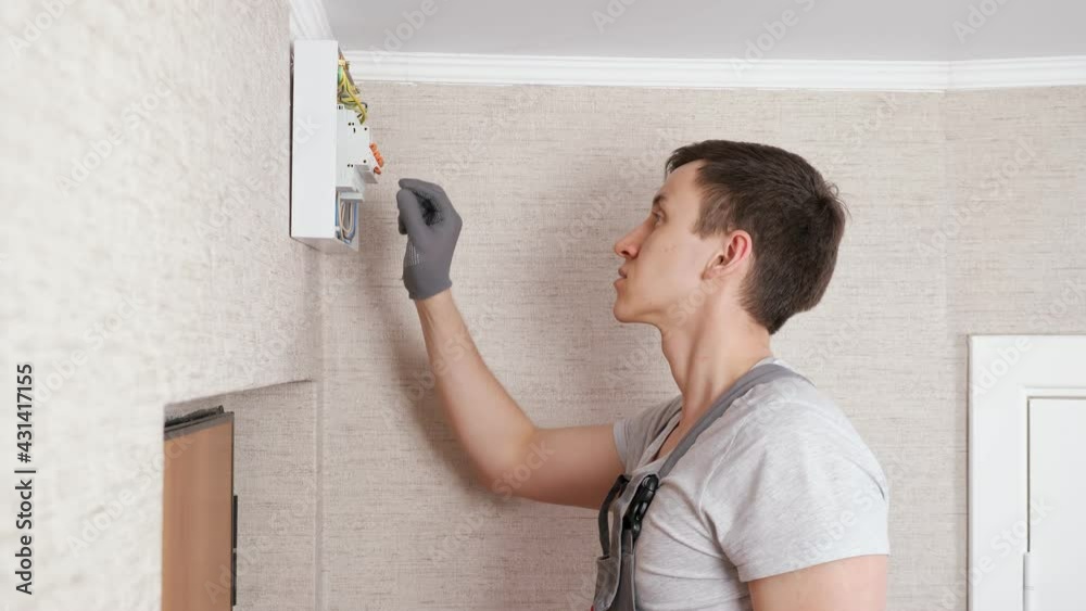 Professional electrician in jumpsuit presses levers on contemporary switchboard to fix above front door in entry room close side view