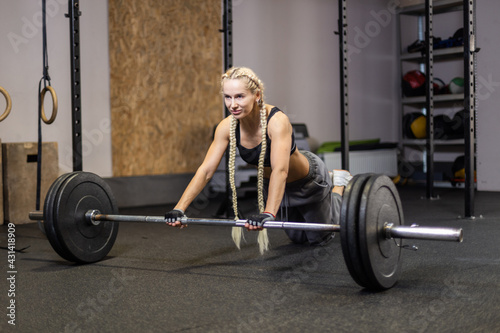 Slim fit woman with long pigtails trains the abdominal muscles, rolls the barbell on the floor in a modern functional gym. Perfect body and trained muscles concept