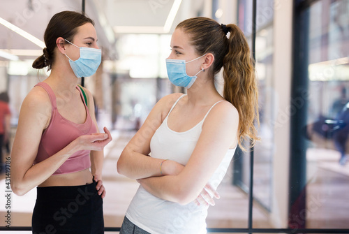 Young girls in sportswear and protective face masks talking friendly in modern fitness studio hall after group pilates workout. New life reality in pandemic