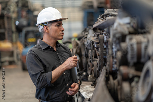 senior engineer using laptop computer checking list old used car engine in machinery parts shop warehouse. old technician man Repair vehicle automobile machine in Recycle motor factory