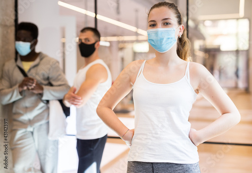 Portrait of confident attractive girl in sportswear and protective face mask standing in modern fitness studio hall before group pilates workout. Active lifestyle and pandemic precautions..