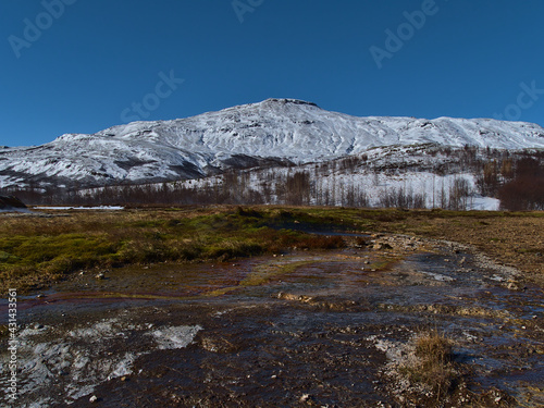 Beautiful view of geothermal area Geysir in Haukadalur, Iceland, part of Golden Circle, in winter season with hot spring, green meadow and snow-covered, rugged mountains on sunny day.