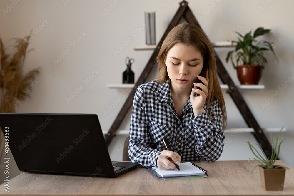 Girl manager of an online store takes an online order by mobile phone while sitting at a table with a laptop at work. The hotline operator writes information in a notebook. Girl manager of an online store takes an online order by mobile phone while sitting at a table with a laptop at work. The hotline operator writes information in a notebook.