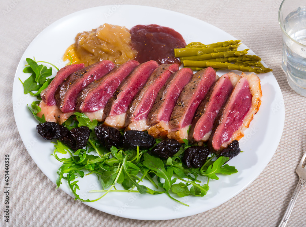 Plate of appetizing roasted duck breast Magret with prunes, sauces and greens