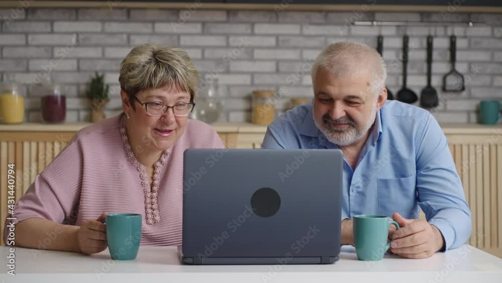 elderly man and woman are drinking tea at home kitchen and viewing movie or pictures on laptop screen