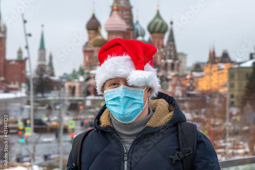 Portrait of caucasian man wearing blue protective face mask and red Santa hat. Blurred Saint Basil's Cathedral and Moscow Kremlin in the background. Theme of Christmas in Russia during coronavirus.