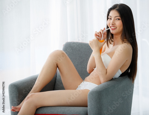 Beautiful young naughty sexy girl on white underwear resting on grey sofa in bedroom and happily drink sweet orange juice for good health after wake up in bright morning of rest day