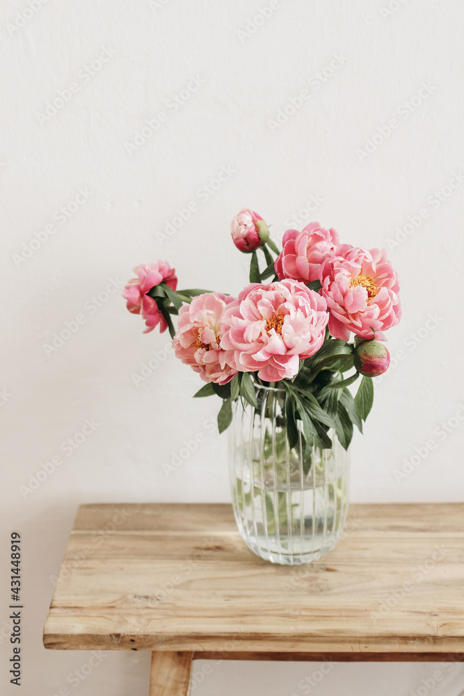 Floral still life scene. Pink peonies flowers, bouquet in glass vase on wooden table. White wall. Selective focus, blurred background. Wedding, birthday celeberation concept. Lifestyle vertical photo