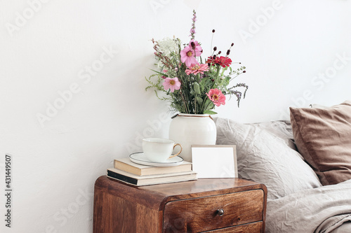 Cup of coffee and books on retro wooden bedside table. Blank greeting card mockup. White ceramic vase. Buquet of cosmos and zinnia flowers. Beige linen, velvet pillows in bed. Scandinavian interior. photo