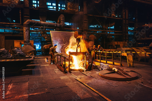 Foundry workshop interior, molten iron pouring from blast furnace into ladle container and workers founders control process, heavy metallurgy industry.