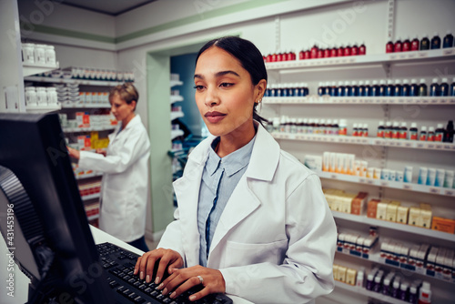 Young woman working in pharmacy typing on computer while checking inventory wearing labcoat standing behind counter