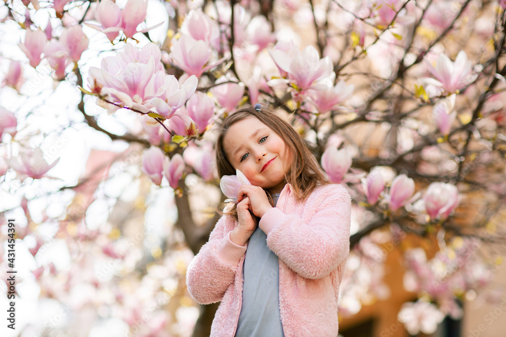 Fototapeta premium Beautiful smiling little girl in the spring garden near blooming magnolia tree
