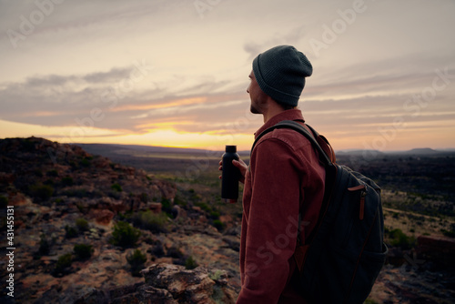 Young male hiker holding water bottle with backpack and cap looking at morning sunrise