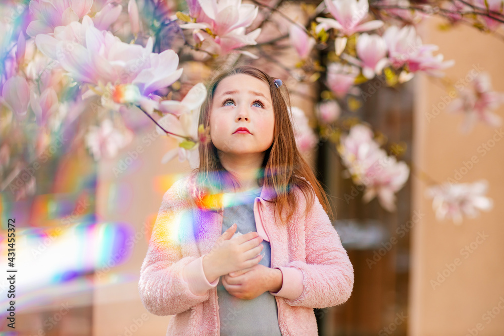 Fototapeta premium Portrait of a cute cheerful girl in a grey dress wearing rosa jacket under a flowering pink magnolia tree