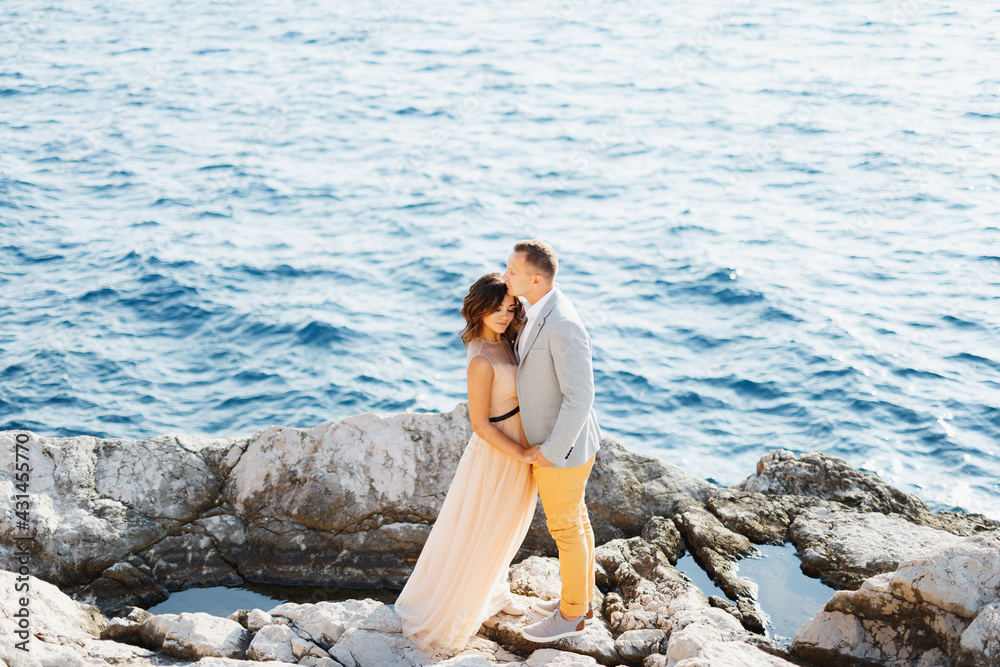 Groom holds the bride's hands while standing on the rocks above the blue sea. View from above