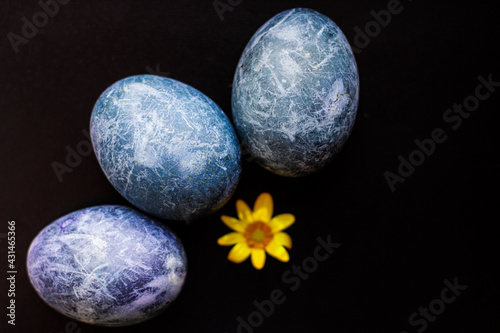 Three painted Easter eggs in the form of blue planets painted in hibiscus on a black background with a yellow flower nearby