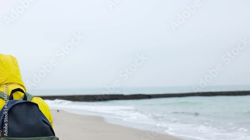 Adult woman in warm yellow jacket with a backpack uses a protective medical mask, walking walks along the seashore and enjoying beautiful view of the sea. Global image of the COVID-19 pandemic concept photo