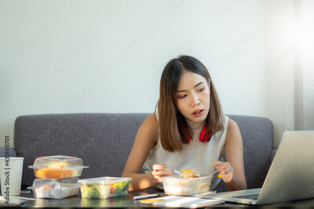 Portrait of young Asian woman freelancer working with laptop at her home and eating Asian food with takeaway. Work at home during self-isolation quarantine and New normal concept. Portrait of young Asian woman freelancer working with laptop at her home and eating Asian food with takeaway. Work at home during self-isolation quarantine and New normal concept.