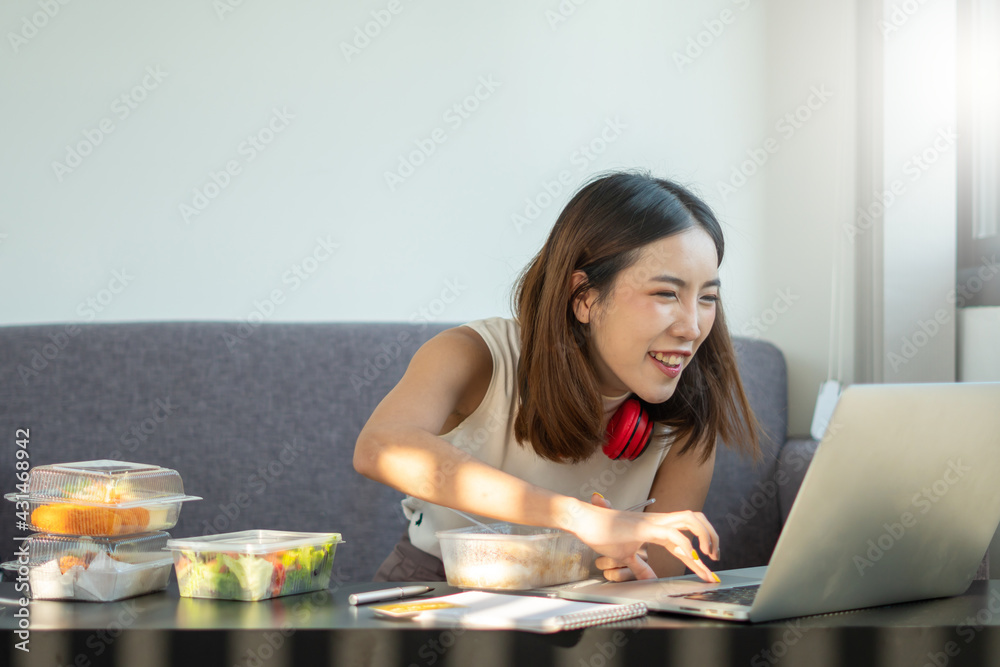 Portrait of young Asian woman freelancer working with laptop at her home and eating Asian food with takeaway. Work at home during self-isolation quarantine and New normal concept. Portrait of young Asian woman freelancer working with laptop at her home and eating Asian food with takeaway. Work at home during self-isolation quarantine and New normal concept.